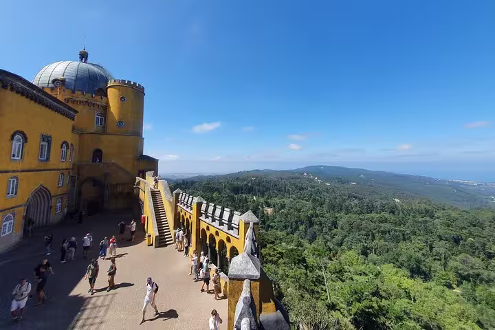 Tourists explore the vibrant Pena Palace in Sintra with panoramic views of lush forests on a sunny day during a Lisbon tour.