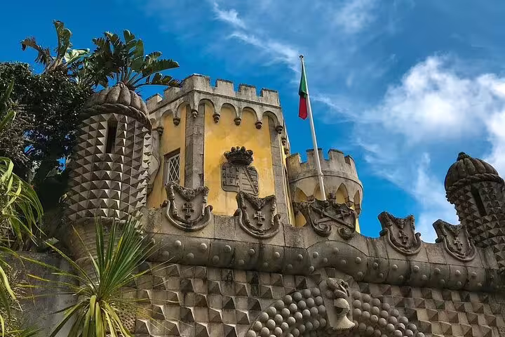 Pena Palace's ornate battlements and vibrant facade under a clear blue sky, featuring lush greenery and a Portuguese flag.