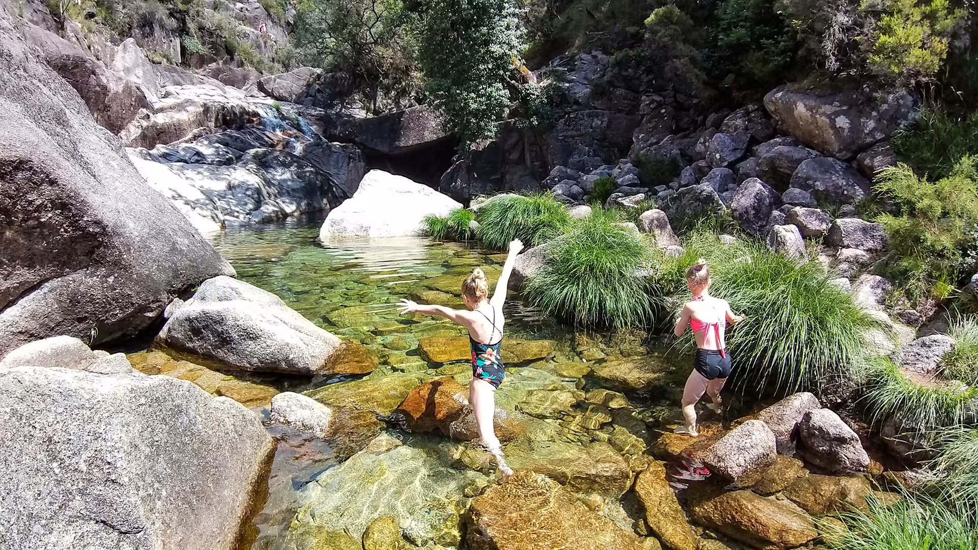 Two women explore a crystal-clear stream surrounded by lush greenery and rocks in Peneda-Gerês National Park.