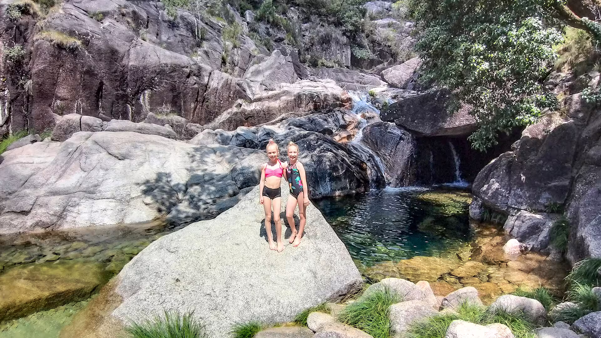 Two children enjoy a sunny day by a serene waterfall in Peneda-Gerês National Park, perfect for nature enthusiasts.