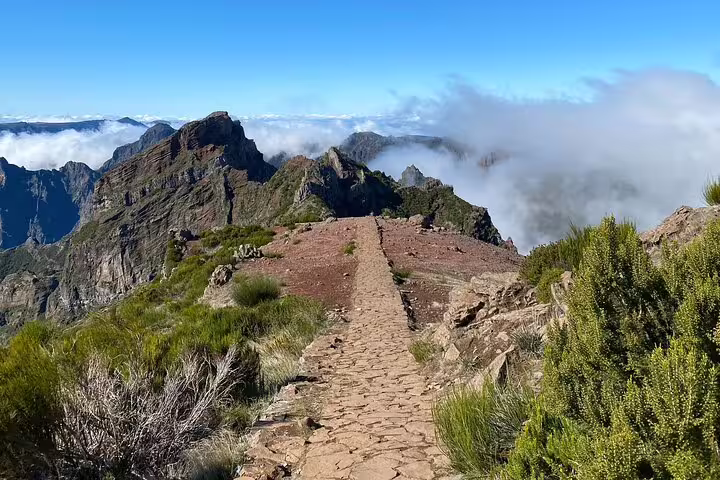 A scenic hiking trail leading to Pico do Arieiro, surrounded by dramatic cliffs and clouds in the background.