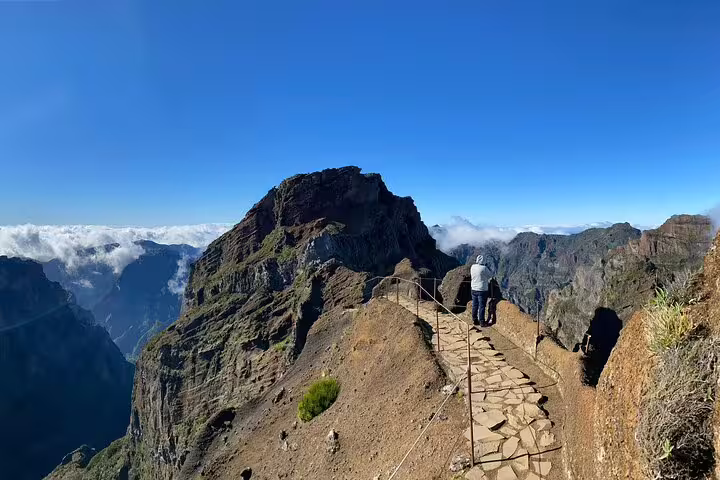 Hikers walk along a narrow path with panoramic mountain views and clear blue skies on Pico do Arieiro trail.