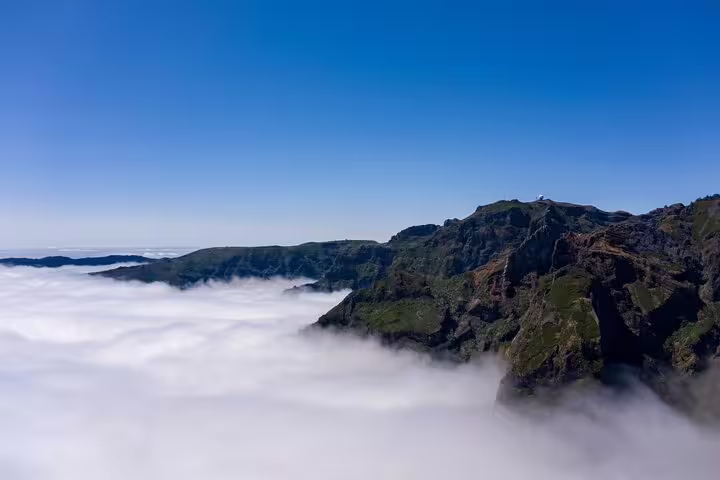 A breathtaking view of Pico do Arieiro with lush cliffs rising above a sea of clouds under a clear blue sky.