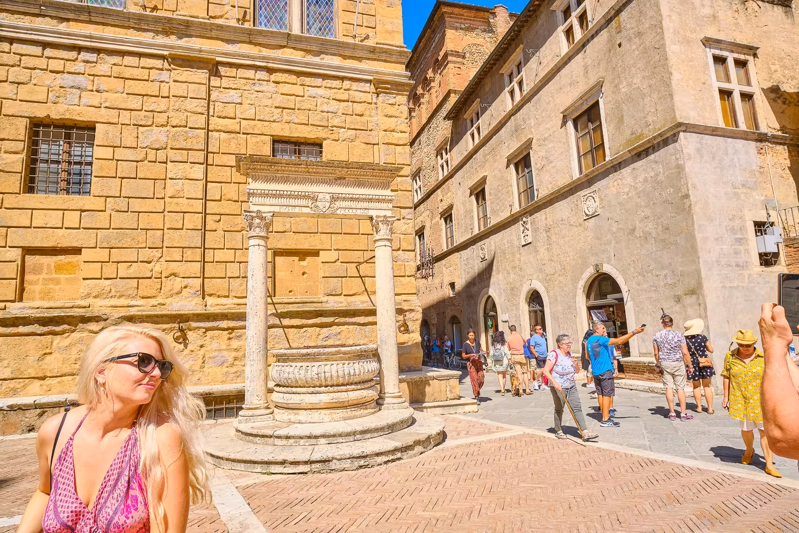 Visitors enjoy the sunlit streets of Pienza, Tuscany, showcasing a charming well and historic stone buildings.