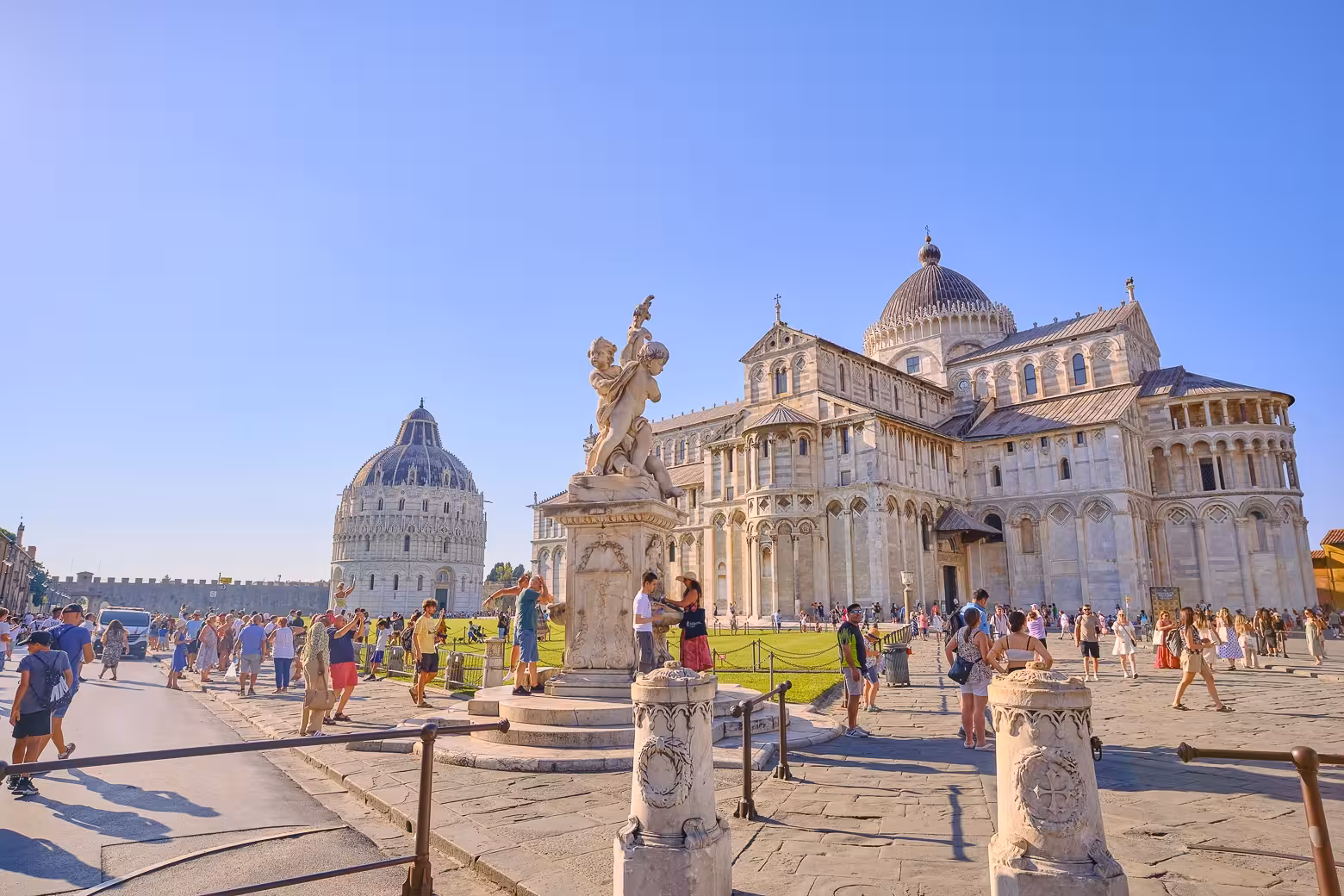 Tourists exploring Pisa's Cathedral Square with the iconic Leaning Tower and Baptistery under a clear blue sky.