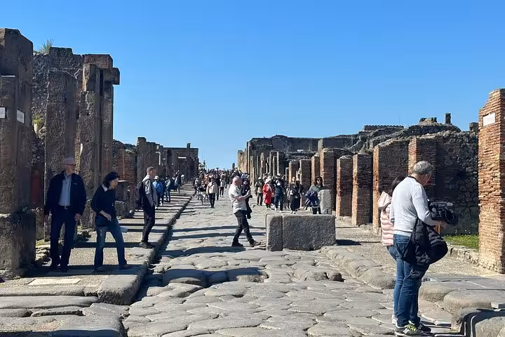 Tourists exploring the ancient stone streets of Pompeii under a clear blue sky, immersed in historical ruins.