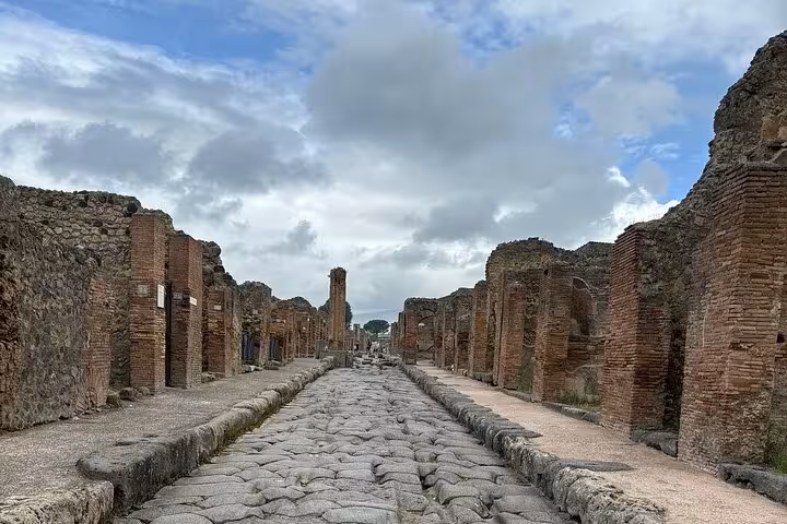 View of a stone-paved street lined with ancient brick structures in Pompeii, under a partly cloudy sky.