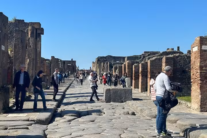Tourists exploring the well-preserved stone streets of Pompeii with ancient ruins lining the pathway under a clear blue sky.