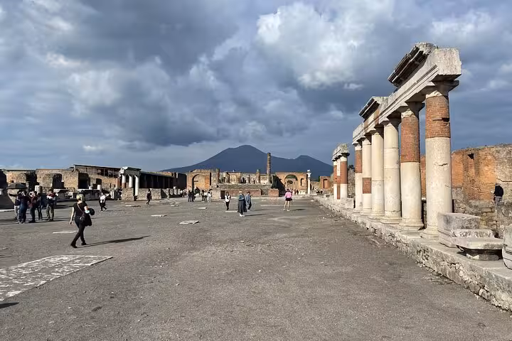 Tourists walk through Pompeii's historic ruins under a dramatic cloudy sky, with Mount Vesuvius visible.