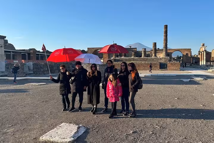 Tour group with red umbrellas exploring ancient ruins in Pompeii with stunning architecture and Mount Vesuvius backdrop.