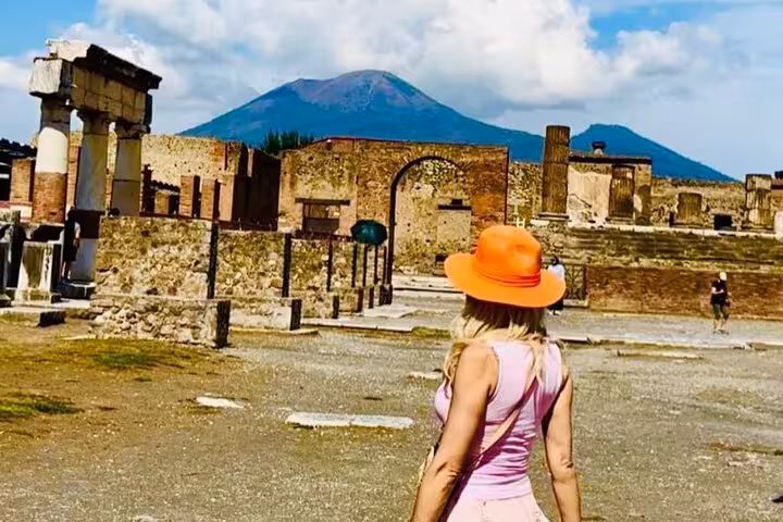 Visitor in orange hat explores ancient Pompeii ruins with Mount Vesuvius in the background on a sunny day tour.