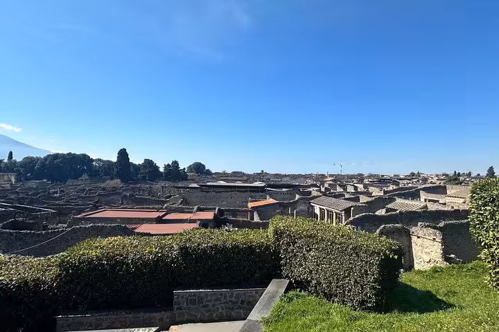 Panoramic view of Pompeii ruins under clear blue skies, showcasing ancient architecture and lush greenery near Vesuvius.