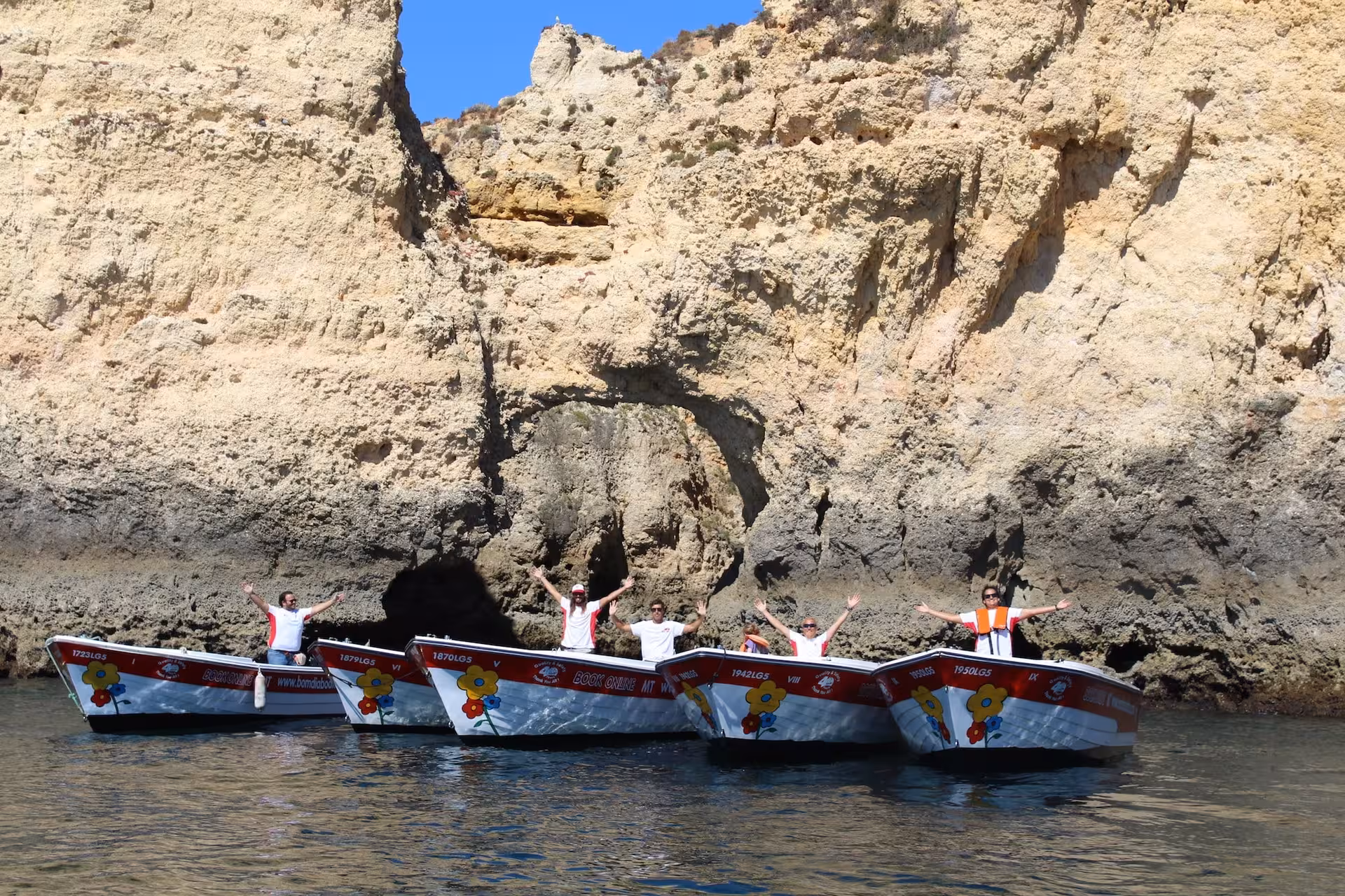 Colorful boats with smiling crew line up beneath towering golden cliffs on a sunny Ponta da Piedade grotto tour in Lagos
