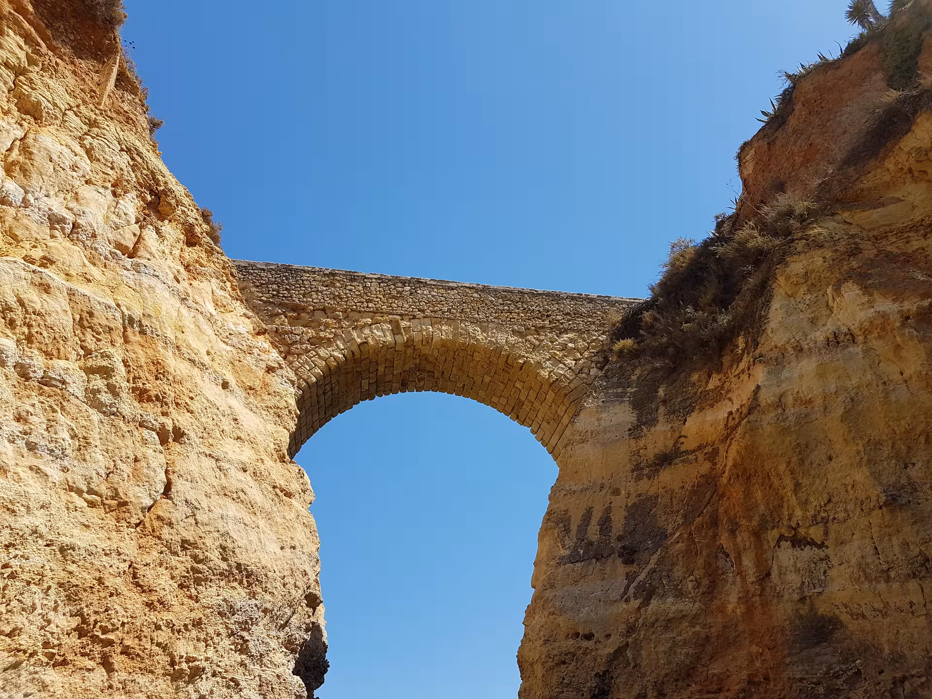Stone arch bridge spanning rugged golden cliffs under a clear blue sky at Ponta da Piedade coastal viewpoint in Lagos
