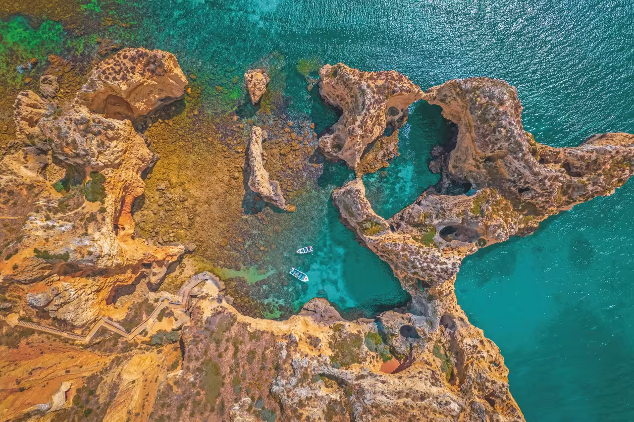 Aerial view of two skiff boats exploring the turquoise waters and dramatic rock arches of Ponta da Piedade coastline in Lagos, Portugal.