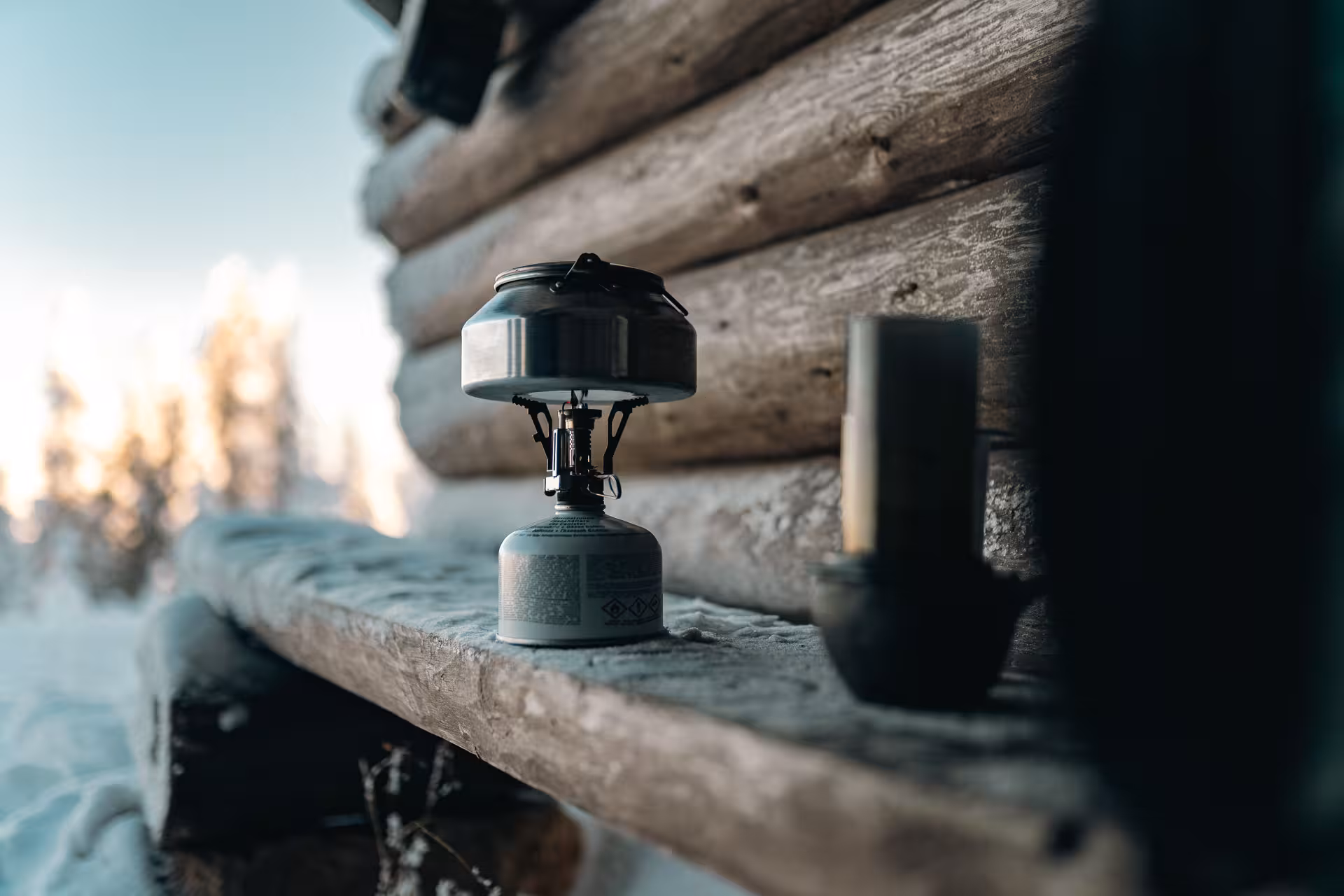 Portable camping stove on a wooden bench outside a rustic cabin in snowy Rovaniemi, Arctic wilderness setting.