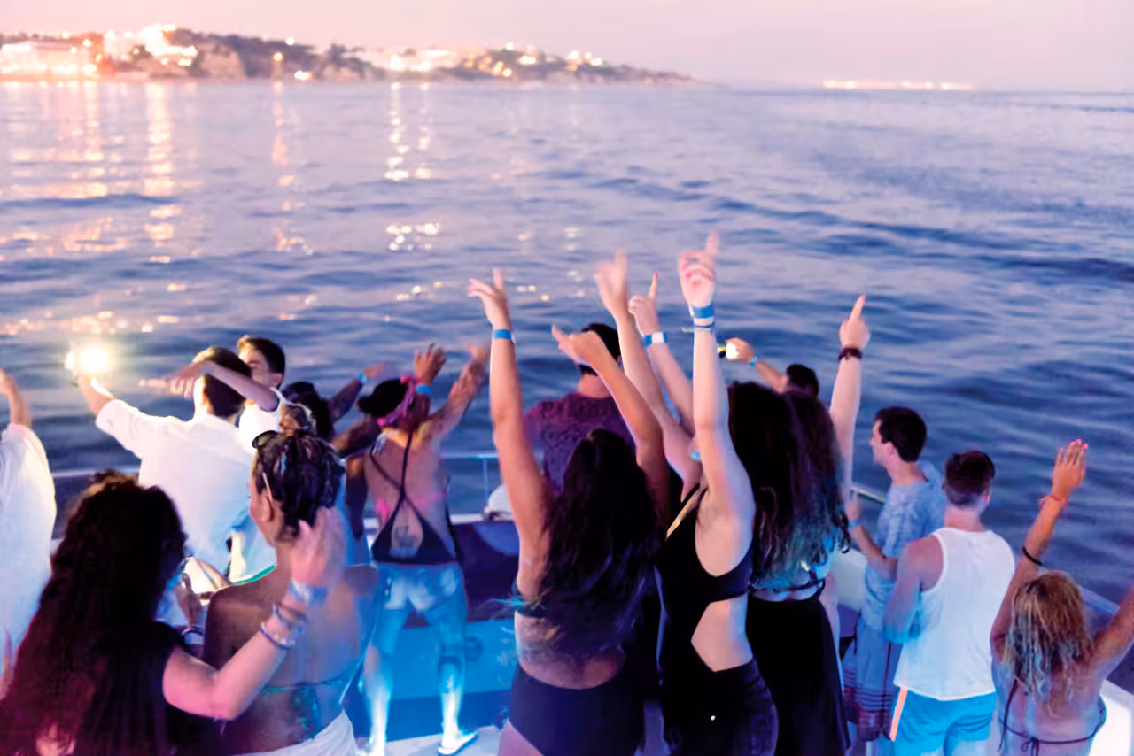 Energetic group dancing with hands raised on a Portimao evening boat party with lit coastline backdrop.