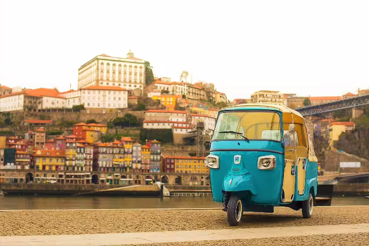 Blue tuk tuk parked along the scenic Douro River with colorful Porto hillside buildings and historic architecture in the background.