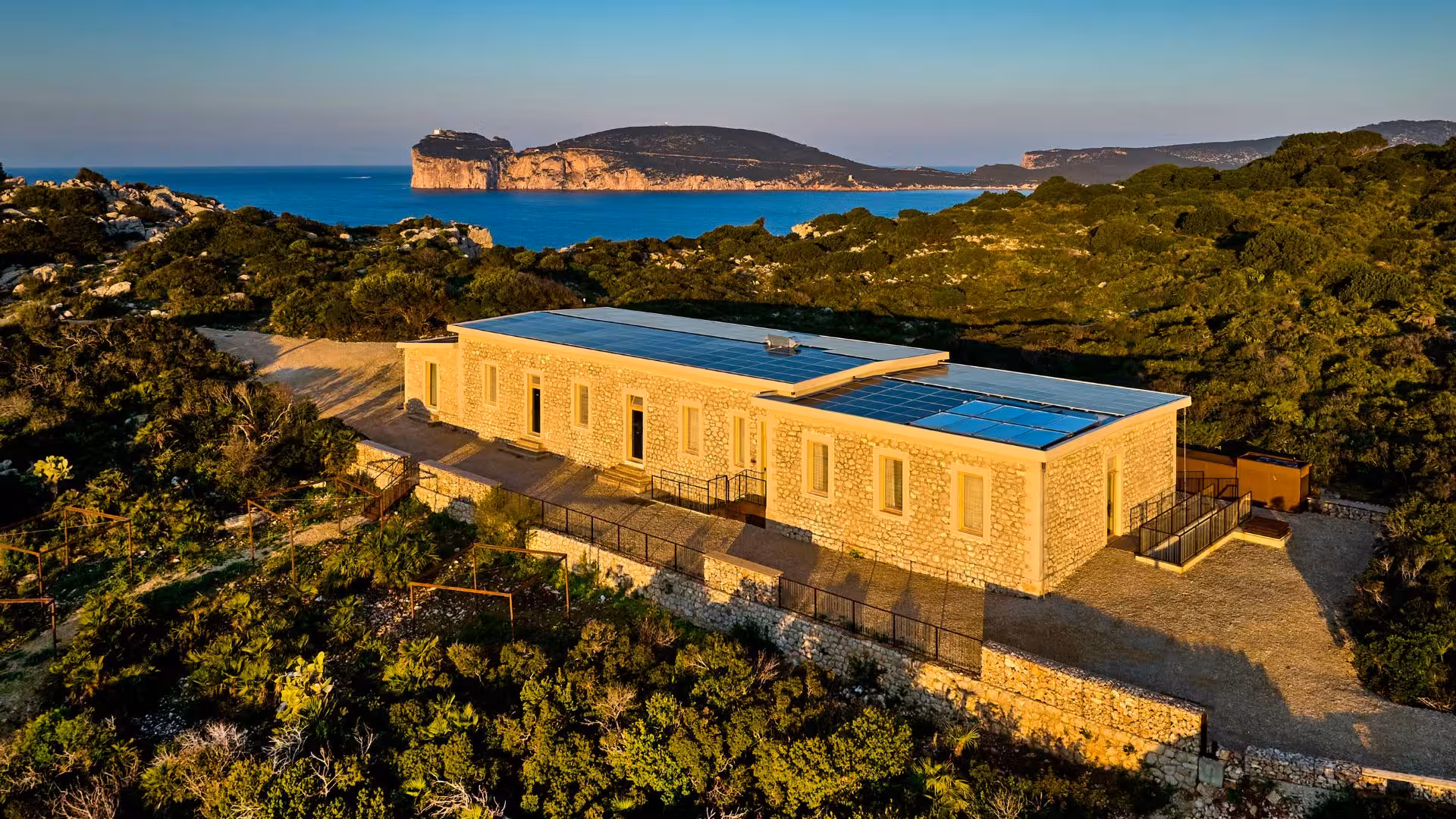 Aerial view of a solar-powered building surrounded by lush greenery and coastal cliffs in Porto Conte, Alghero.
