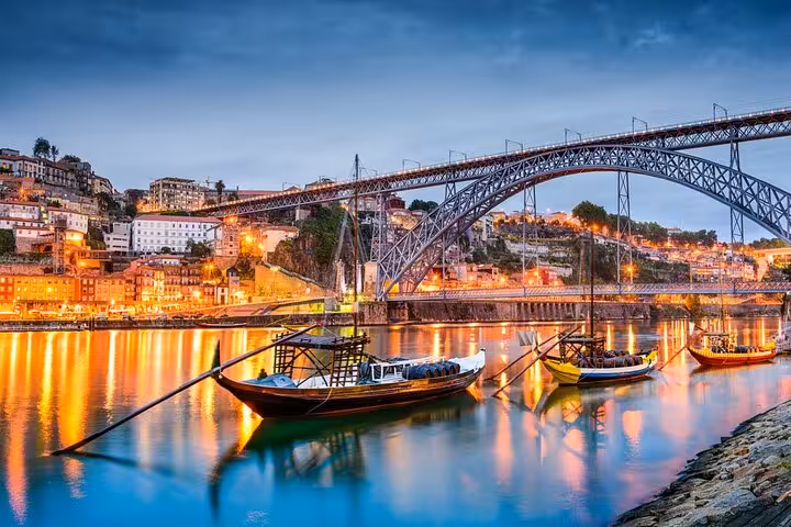 Scenic evening view of Porto's Dom Luís I Bridge with traditional boats on the Douro River illuminated by city lights.
