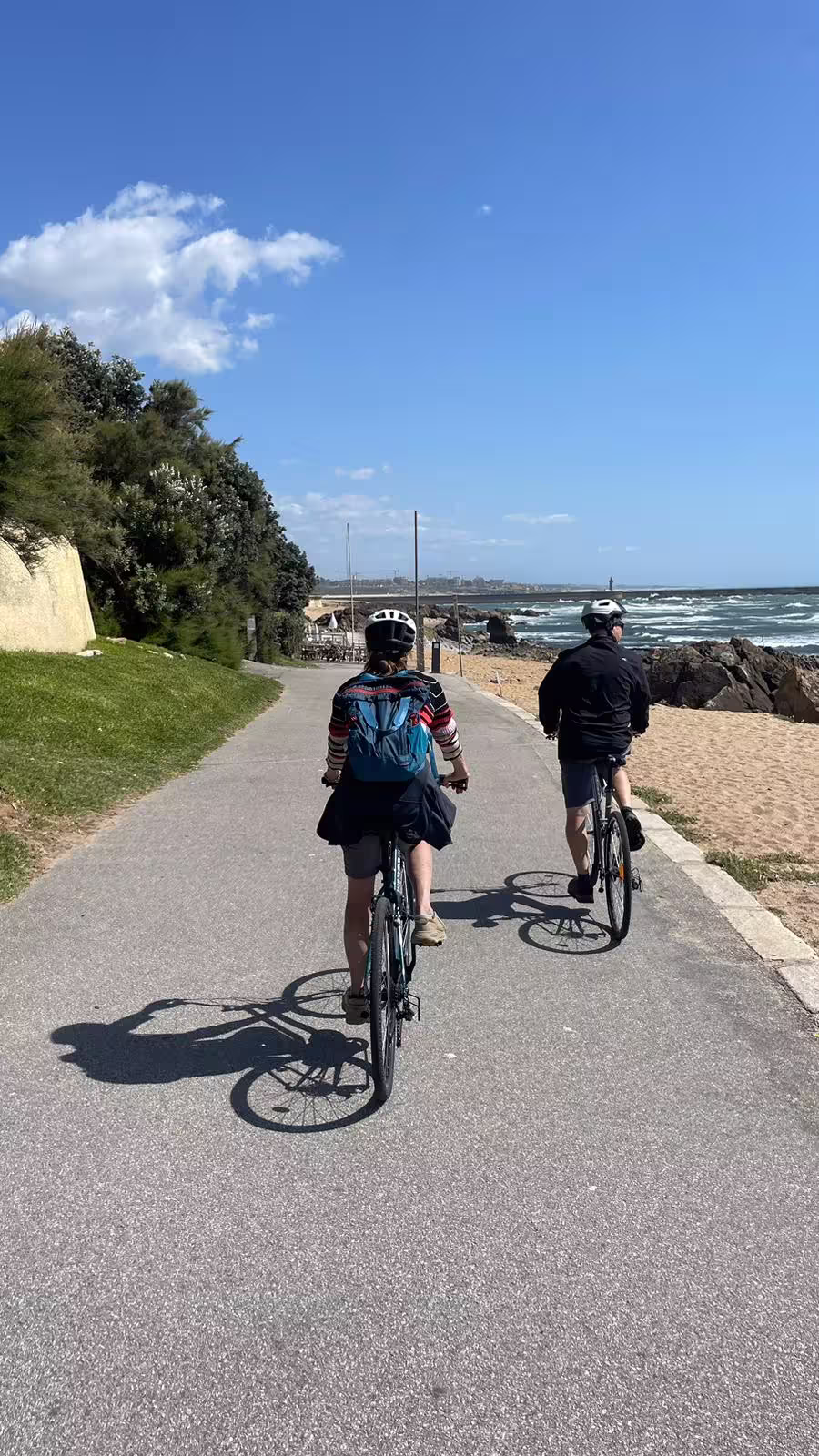 Two cyclists pedal along a picturesque coastal path in Porto, enjoying sunny skies and scenic ocean views on e-bike tour.