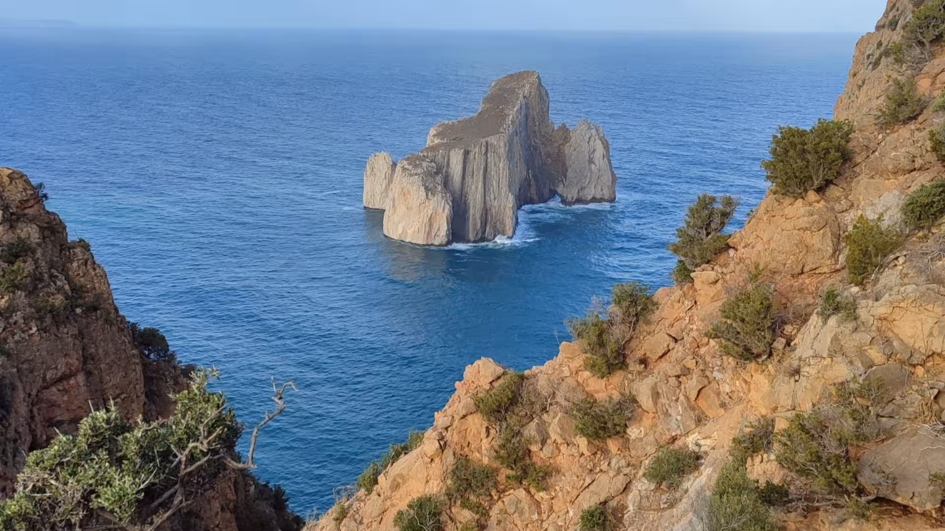 Iconic sea stack emerges from turquoise waters, framed by dramatic cliffs in Porto Flavia canyoning landscape.