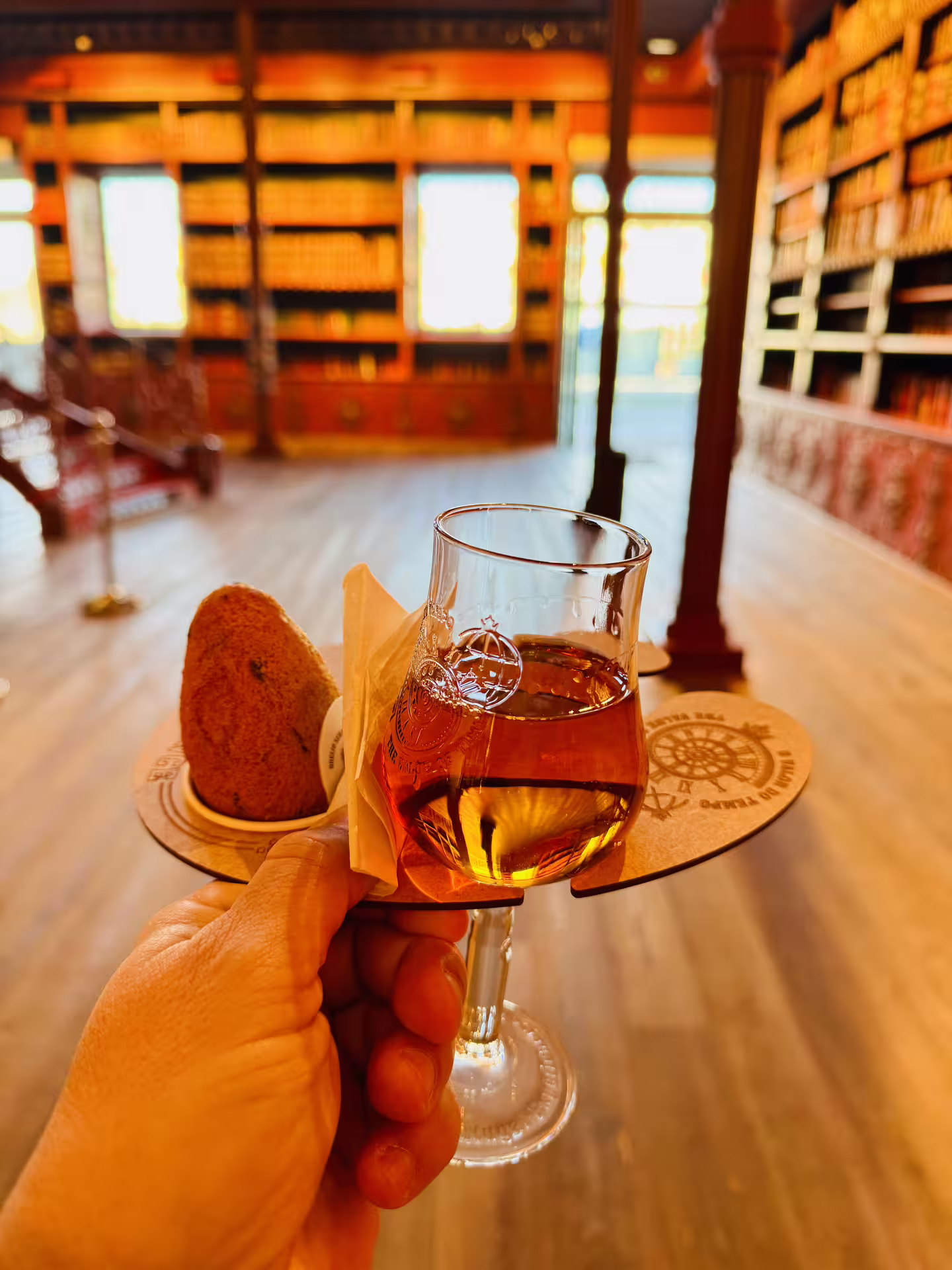 Hand holding a glass of Port wine with a pastry in a historic library setting during a Porto and Gaia walking tour.