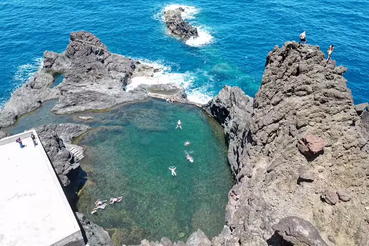 Aerial view of natural volcanic pools in Porto Moniz, Madeira, with tourists swimming and exploring rugged coastline on a sunny day.