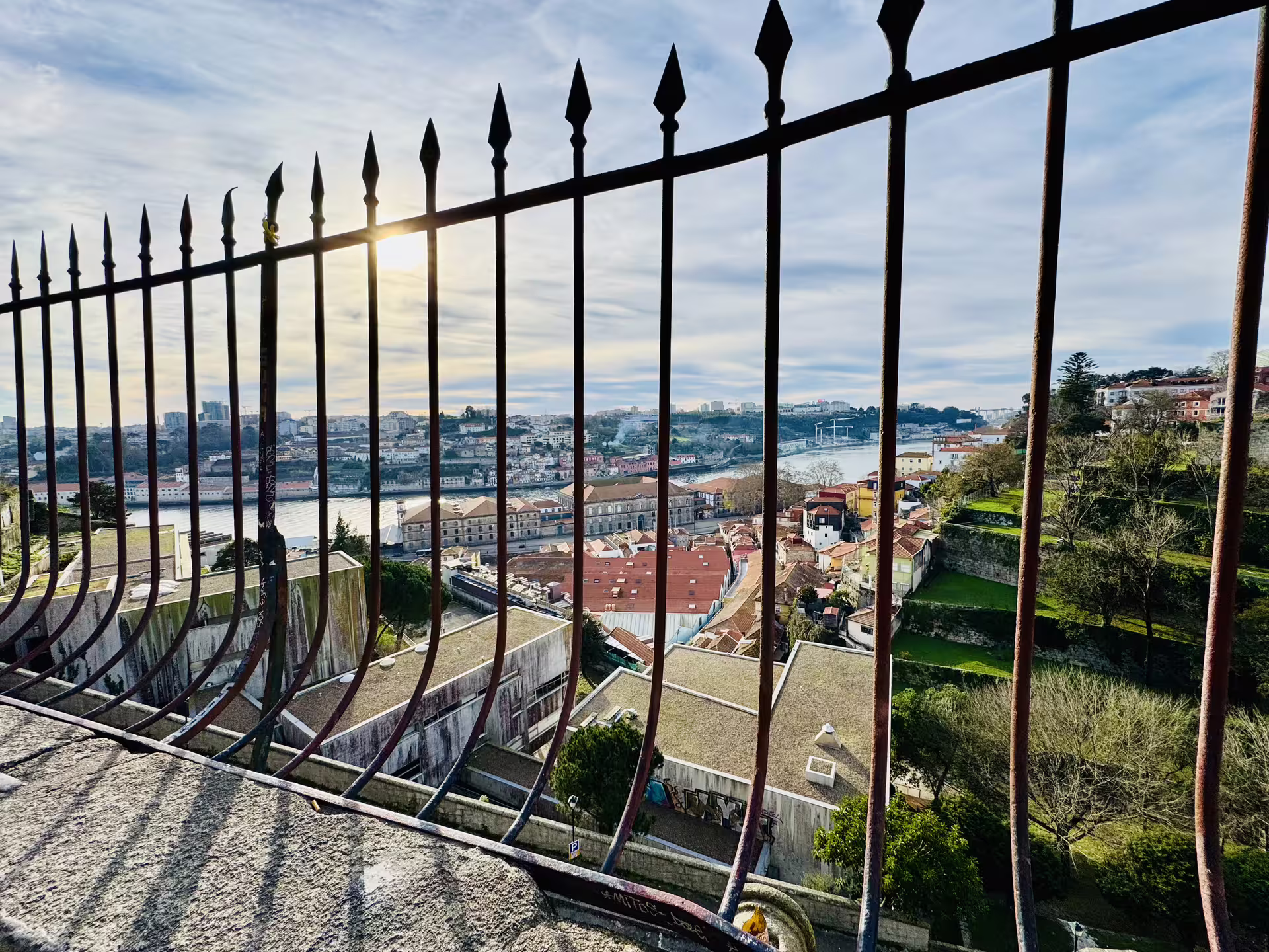 Scenic view of Porto's old town and Douro River from behind a wrought iron fence, highlighting historic architecture and greenery.