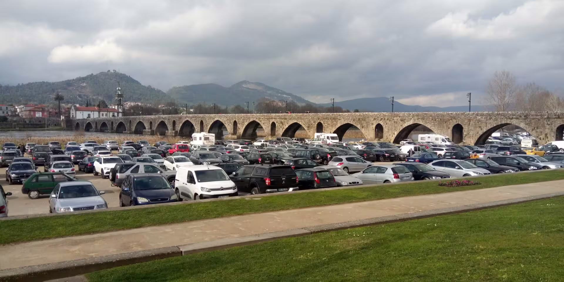 Parking area full of cars with scenic view of historical Roman bridge in Pontevedra, Spain, under cloudy skies.