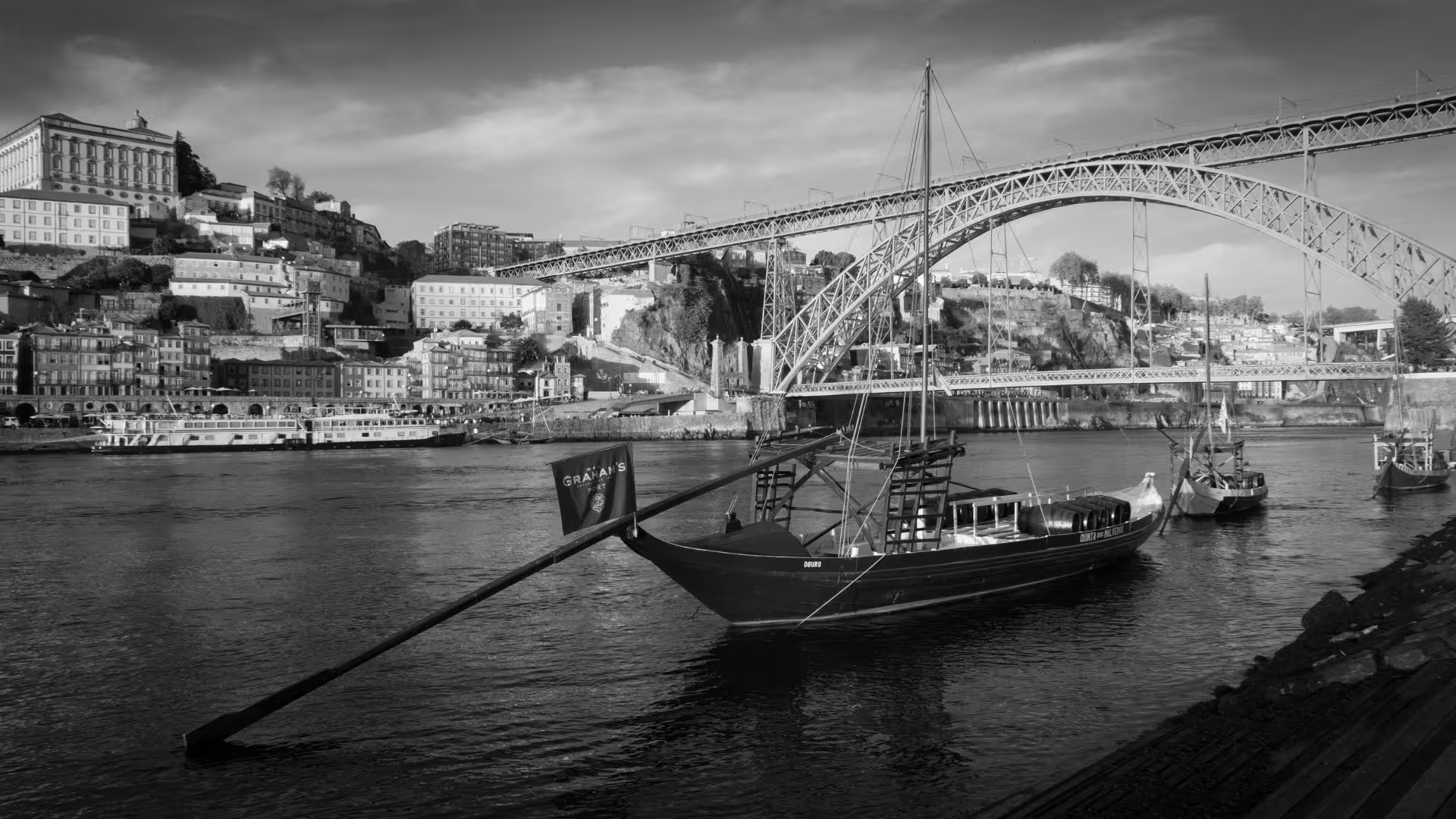Traditional rabelo boats float on the Douro River with Porto's historic Ribeira district and Dom Luís I Bridge in view.