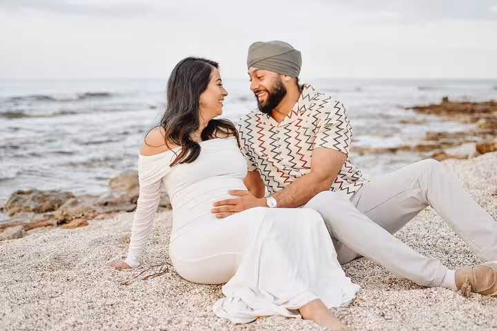 Couple sitting on Heraklion beach, woman in white maternity dress and man in casual attire, sharing a joyful moment by the sea.