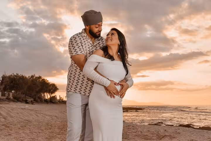 Couple embracing on Heraklion beach at sunset, woman in white dress, capturing tender pregnancy moment with scenic backdrop.