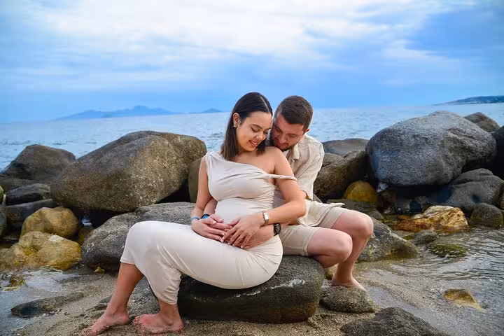 Pregnant couple embracing on Ammoudara beach rocks, enjoying a romantic private photoshoot by the ocean.