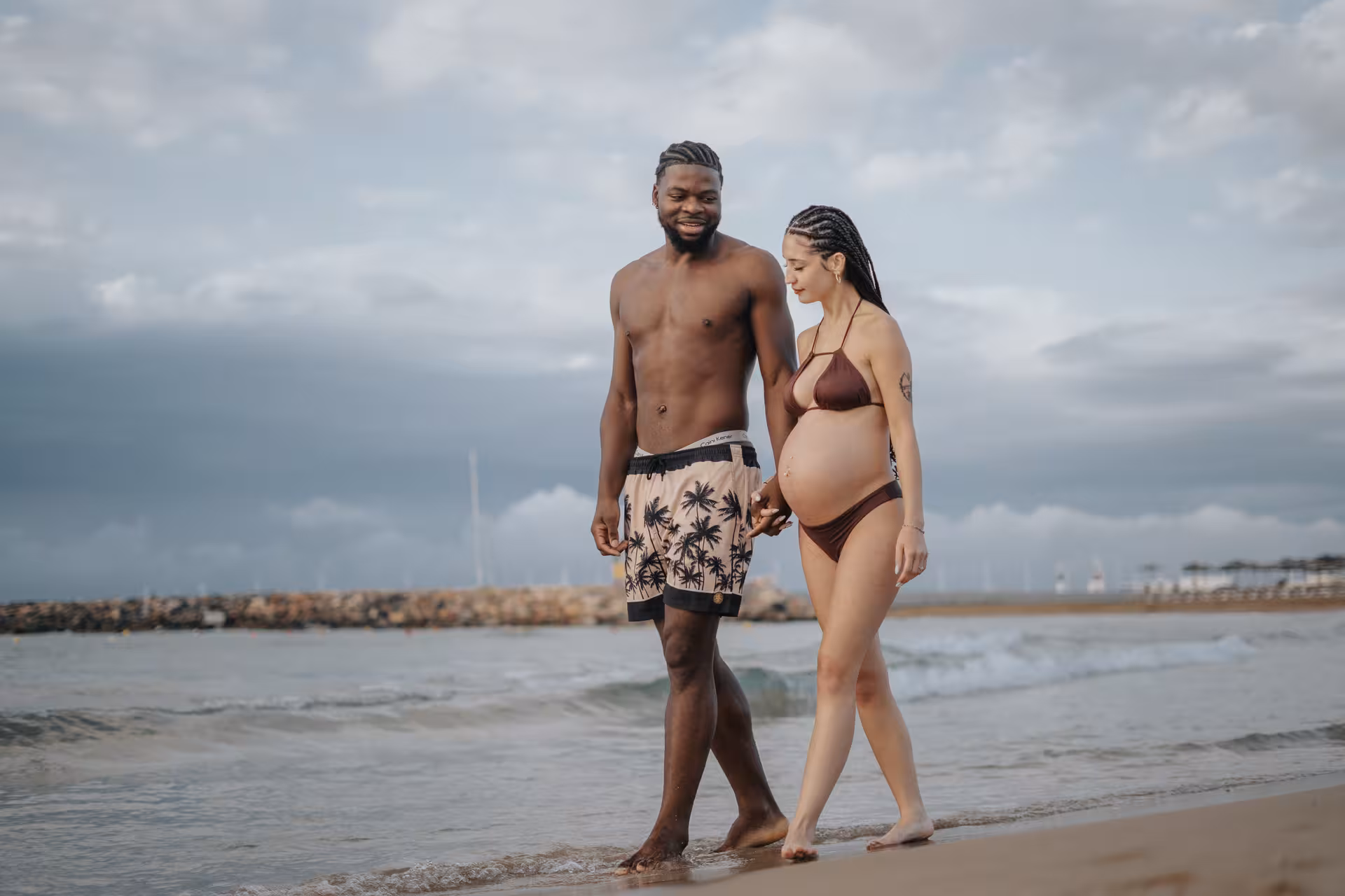Pregnant couple walking hand in hand along Heraklion Venetian Port beach, enjoying a serene photoshoot moment.