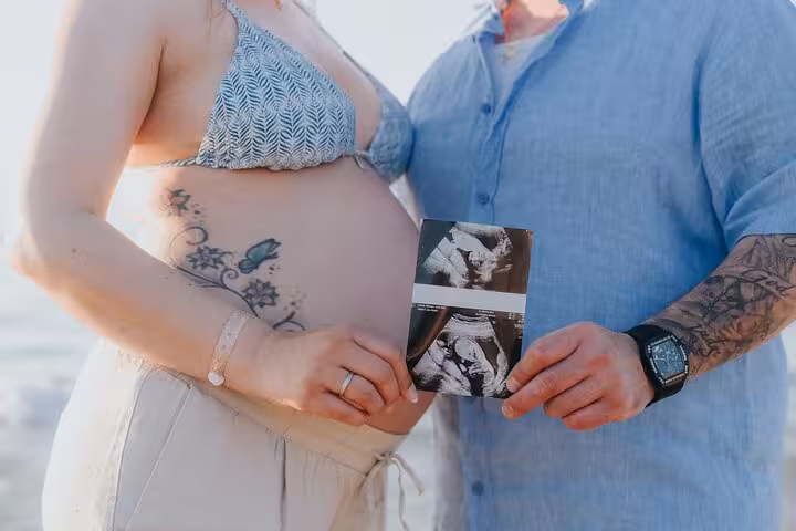 Pregnant couple holding ultrasound photo on Heraklion beach, capturing a precious maternity moment in the sun.