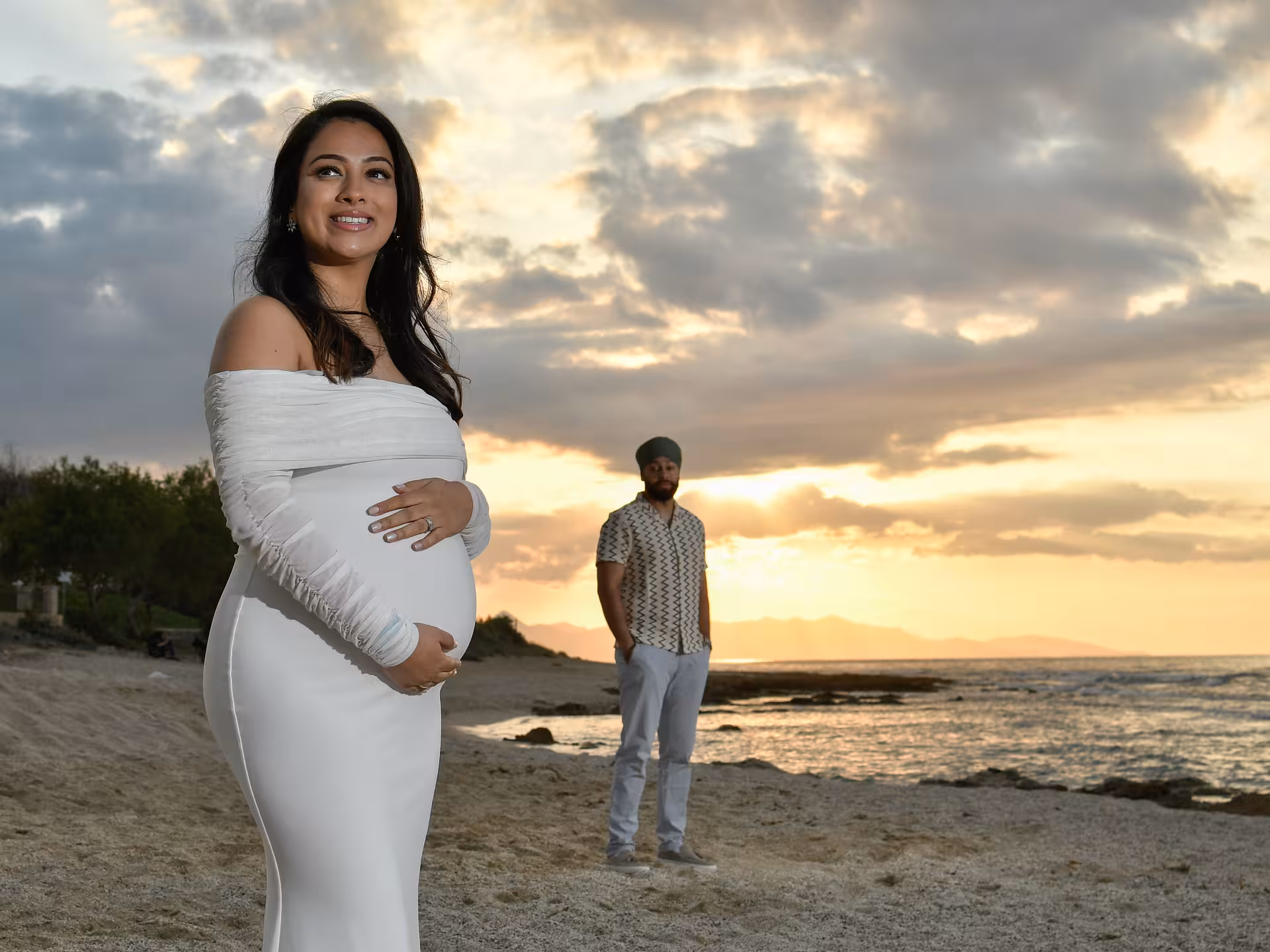 Pregnant woman in white dress on Heraklion beach at sunset with partner in background during private photoshoot.