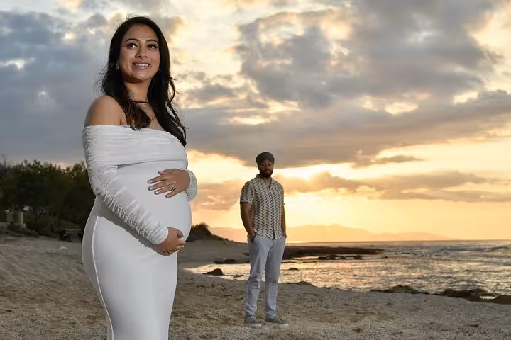 Pregnant woman in white dress on Heraklion beach at sunset, smiling with partner in background, ideal for maternity photoshoot.