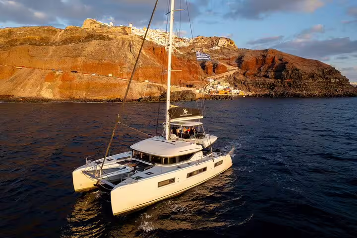 Luxurious catamaran sailing near the rocky coast of Santorini at sunset, offering premium cruise experiences.