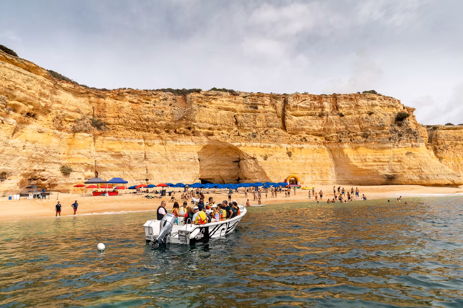 Private boat approaches a scenic Algarve beach with vibrant umbrellas and limestone cliffs in the background.