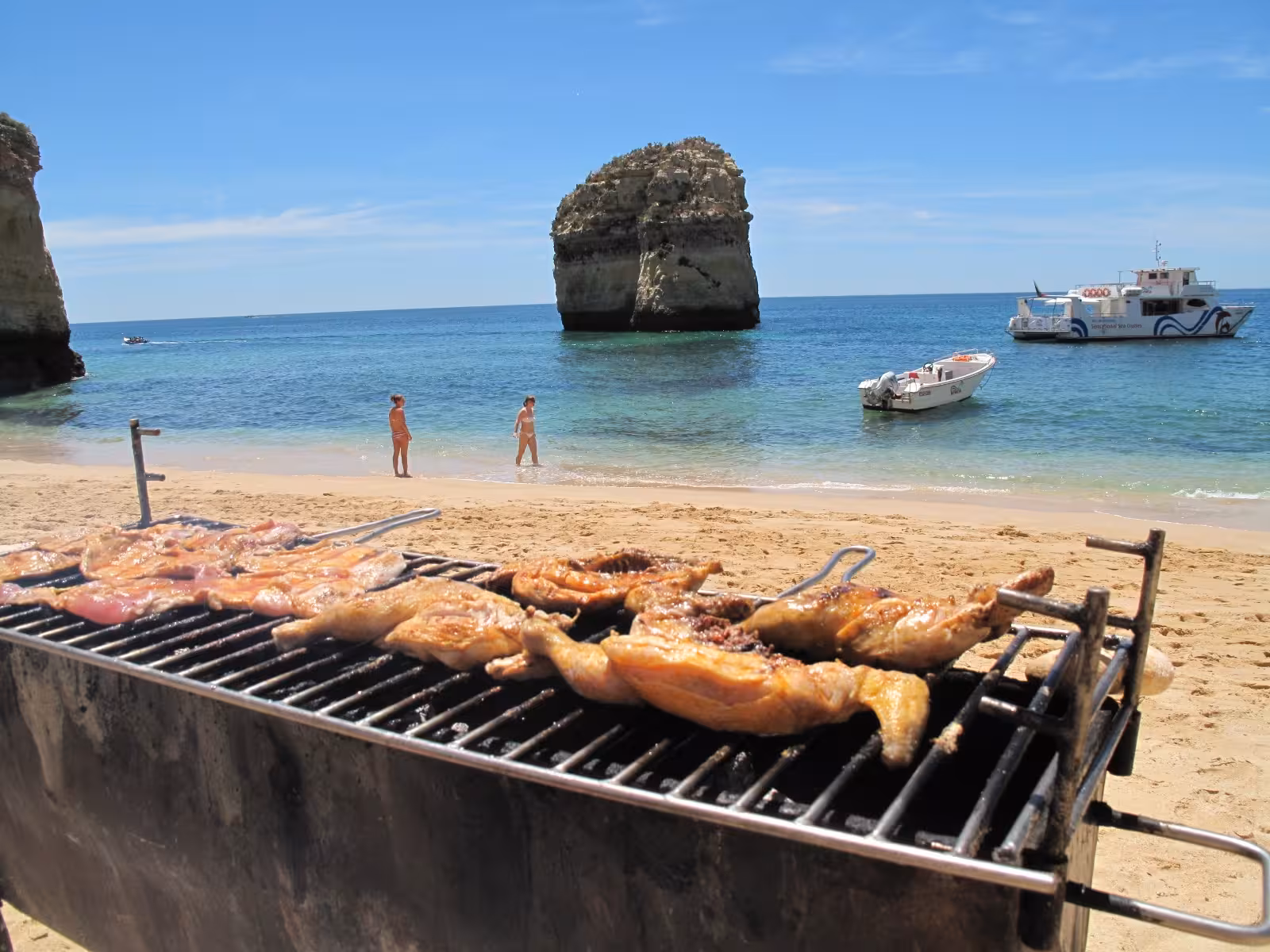 Beachfront barbecue with sizzling meats and anchored private boats against the backdrop of Algarve's azure waters.