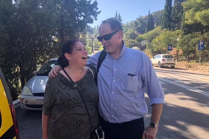 Two happy travelers enjoying a sunny day during a private tour from Athens to Delphi, surrounded by lush greenery and cars.