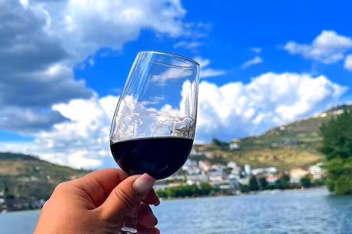 Hand holding a glass of Port wine on a private Douro River boat trip, with hills and riverside village views