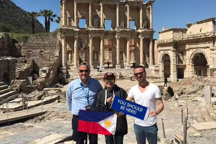 Tourists holding a 'You Should Be Here' sign at Ephesus' Library of Celsus on a private tour from Kusadasi Port.