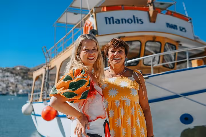 Two women smiling in front of a boat named Manolis, enjoying a private photoshoot experience in Elounda.