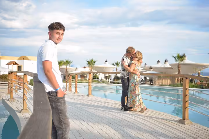 Young man poses by a poolside in Malia while a couple shares a romantic moment in the background during a photoshoot.
