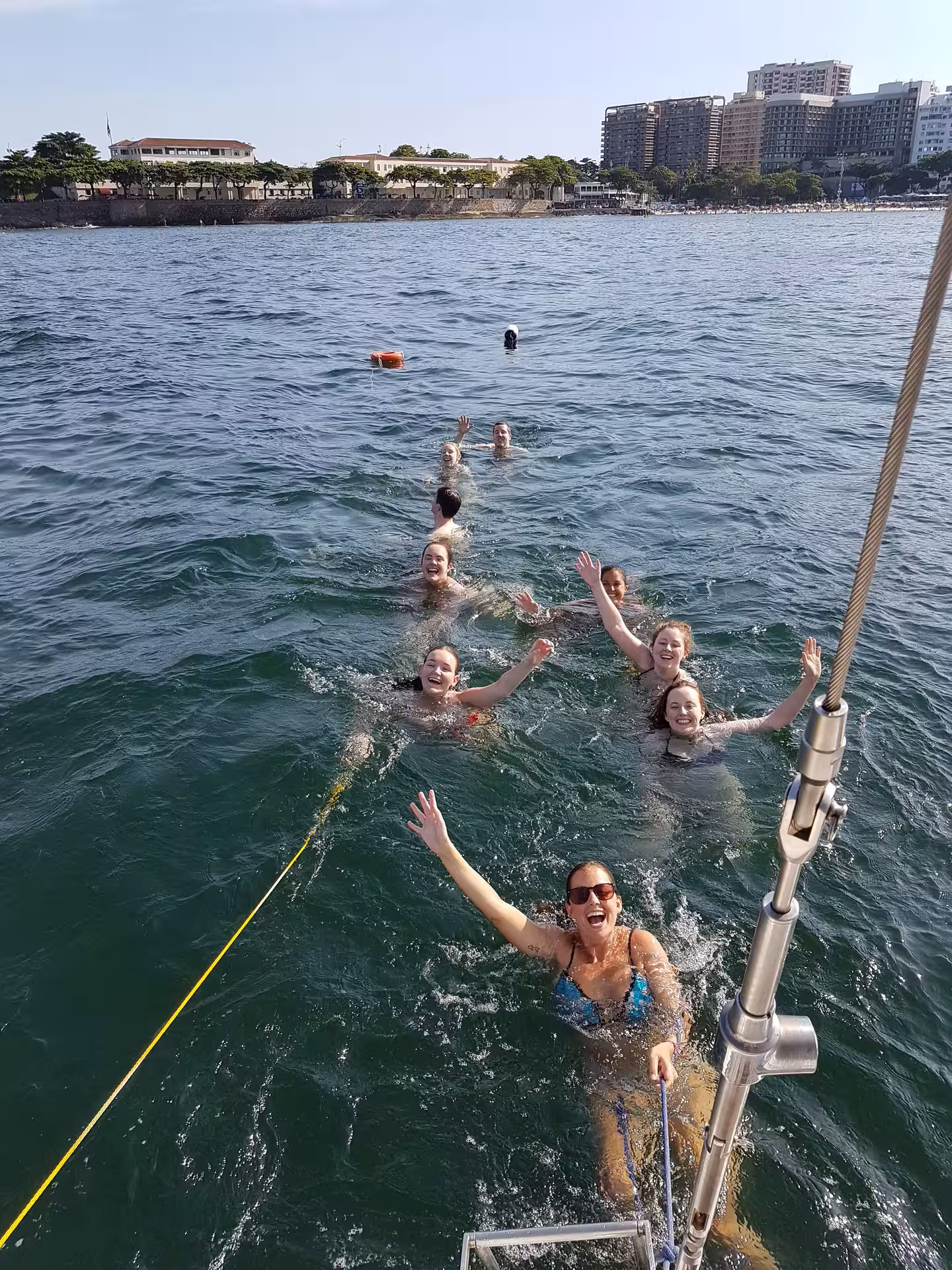 Group enjoying a swim near the sailboat on the Private Sailing Tour KAMEHAMEHA, with city views in the background.