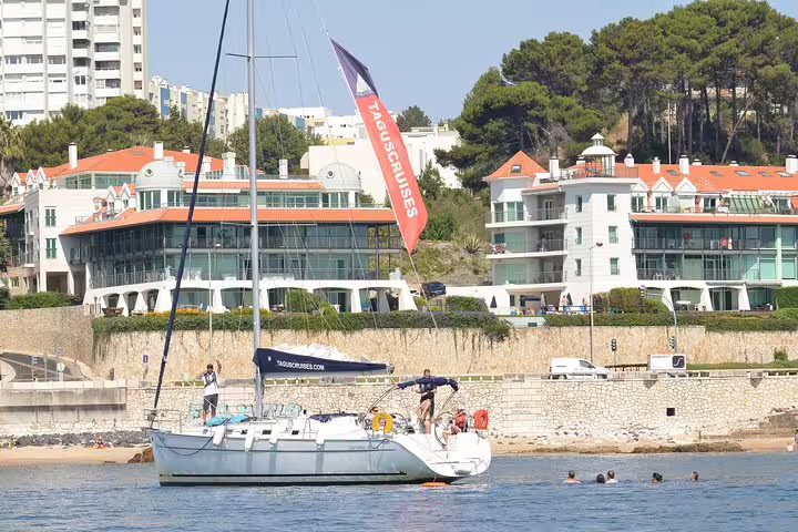 Sailboat anchored near Oeiras beach with people swimming, highlighting picturesque coastal scenery and modern buildings.