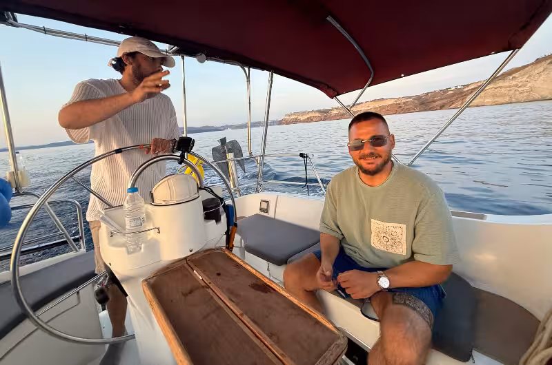 Skipper and guest relaxing on a private Santorini sunset sailing cruise, caldera coastline in background