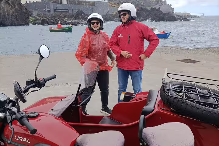 Two people in helmets and rain gear enjoy a sidecar adventure by the seaside in Funchal, highlighting a unique coastal tour experience.